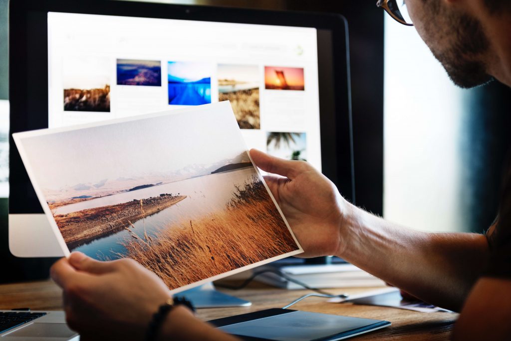 Man holding a photograph while sitting in front of a computer screen featuring an image gallery.