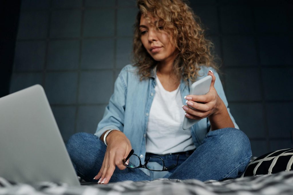 A woman sitting on a bed with a smartphone and laptop.