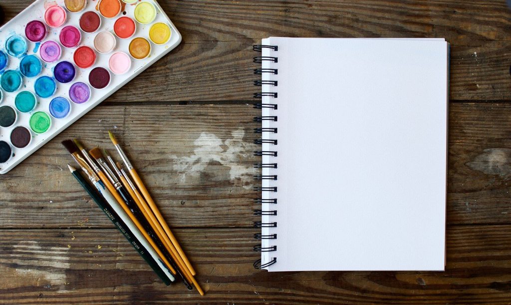 A blank white notebook lying next to a palette of paint and some brushes.