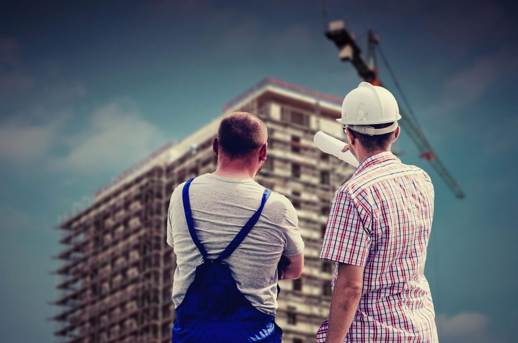Two construction workers examine a building in the distance with their backs to the camera.